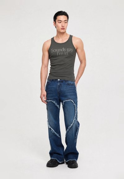 Homme portant un débardeur vert foncé avec le texte "Sounds gay I'm in" et un jean bleu avec des motifs en forme de cœur effilochés, debout devant un fond uni.