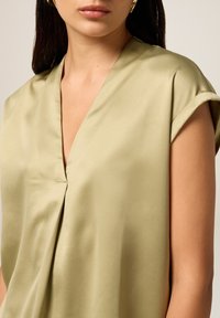 Donna che indossa un top di satin verde oliva chiaro con maniche corte arrotolate e scollatura a V, mostrando parte del suo viso e del collo.