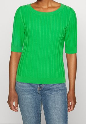 Person iført en lysegrøn, ribstrikket kortærmet sweater sammen med blå denimjeans, stående mod en ensfarvet baggrund.
