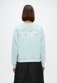 Personne aux cheveux courts et bruns porte un sweat-shirt bleu clair avec "XOXO" et de petites figures colorées dans le dos, associé à un pantalon noir.