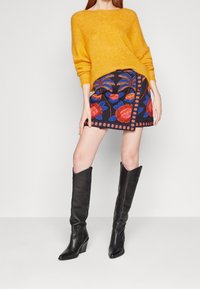 Gul stickad tröja, flerfärgad broderad minikjol med blommönster, och knähöga svarta läderstövlar med klack.