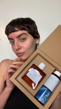 Brązowa szklana butelka z białą nakrętką i etykietą "KYMA", obok niebiesko-białej butelki z wzorem, w kartonowym pudełku trzymanym przez osobę.