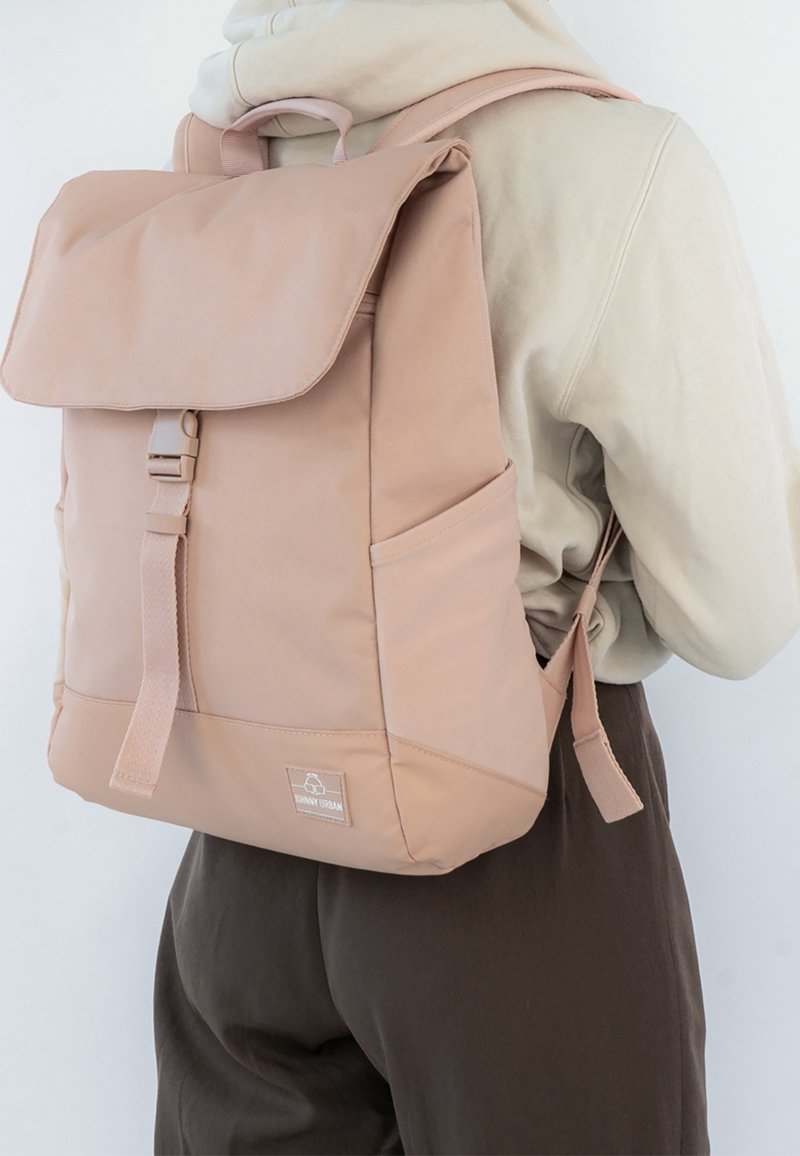 Personne portant une veste beige et un pantalon marron avec un sac à dos rose clair doté d'un rabat supérieur et d'une fermeture à boucle.