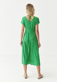 Grünes, strukturiertes Midi-Kleid mit kurzen Ärmeln, tailliertem Schnitt und einem Schlüssel-Ausschnitt hinten, abgerundet mit einer Schleife am Hals.