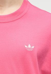 Κοντινό πλάνο ενός ατόμου που φοράει ροζ μπλούζα με στρογγυλή λαιμόκοψη και μικρό λευκό λογότυπο τριφύλλι Adidas στο στήθος.