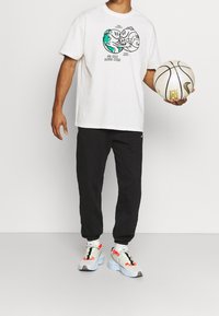 Bílý grafiký tričko s kresleným designem, černé tepláky a vícebarevné tenisky. Osoba drží basketbal.