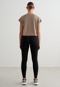 Femme avec un chignon tressé portant un haut taupe à manches courtes, des leggings noirs et des baskets blanches, debout face à un mur blanc uni.