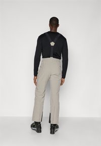 Pantaloni de schi gri deschis cu bretele negre ajustabile, croială taperată și deschideri cu fermoar la glezne, asortați cu cizme negre.