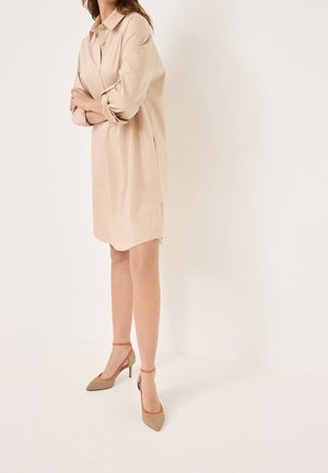 Beżowa sukienka koszulowa z kołnierzykiem, z przodu na guziki i luźnymi rękawami, w połączeniu z jasnobrązowymi szpilkami z czerwonymi paskami na kostkach.