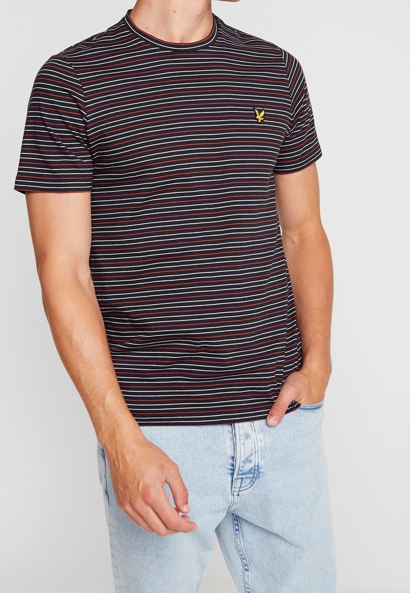Camiseta a rayas en negro, rojo y gris. Cuello redondo, mangas cortas, diseño ajustado. Presenta un pequeño logo de águila amarilla en el pecho.