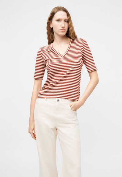 Γυναίκα με κυματιστά κόκκινα μαλλιά που φοράει κοντομάνικη μπλούζα με V λαιμόκοψη και ρίγες κόκκινες και λευκές, και παντελόνι ψηλόμεσο λευκό, με το χέρι στην τσέπη.