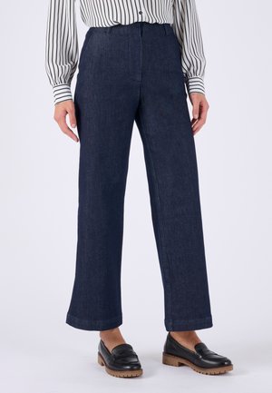 Donna che indossa pantaloni larghi blu scuro, mocassini neri con suole marroni e una camicetta bianca con righe verticali nere.