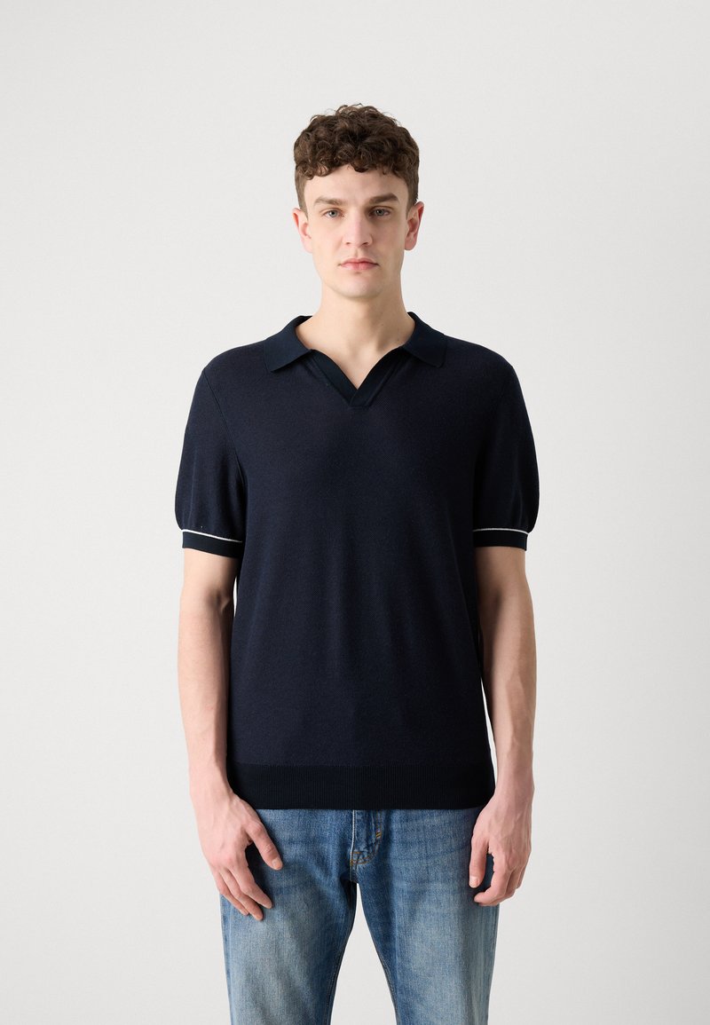 Polo en tricot navy avec un col en V, des manches courtes bouffantes et des poignets texturés contrastants. Tissu doux et détail de bas en côte.