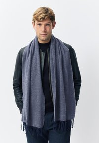 Grauer Wollschal mit weicher Textur, versehen mit einem Fischgrätmuster und fransigen Enden. Über einem schwarzen Oberteil und einer Lederjacke getragen.