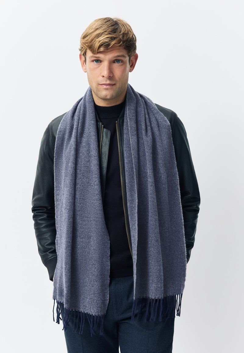 Grauer Wollschal mit weicher Textur, versehen mit einem Fischgrätmuster und fransigen Enden. Über einem schwarzen Oberteil und einer Lederjacke getragen.