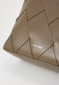 Ruskea kudottu tekonahkainen laukku, jossa on sileä pinta, jossa on valkoinen "Calvin Klein" -logo ja kontrastiväriset tikkausreunat.