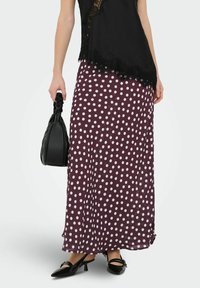 Jupe maxi en bordeaux profond à pois blancs, confectionnée dans un tissu léger. Associée à un haut noir et un sac à main, présentant une texture lisse.