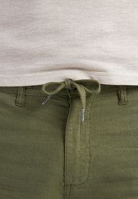 Olive green linen pants with a drawstring closure, featuring a flat front and side pockets, set against a light beige textured shirt.