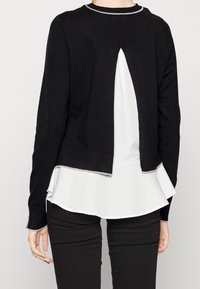 Czarny dzianinowy kardigan z rozcięciem na plecach, wyposażony w biały warstwowy dół peplum i mankiety z srebrnymi akcentami. Dopasowany krój, długie rękawy.