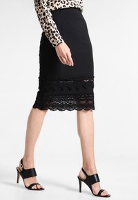 Schwarzer Bleistiftrock mit gestuftem Spitzenbesatz am Saum. Weicher Stoff, tailliertes Design, kombiniert mit schwarzen High-Heel-Sandalen.