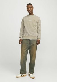 Sudadera gris texturizada con logo blanco en el pecho, combinada con jeans sueltos y desgastados, y zapatillas blancas con acentos verdes.