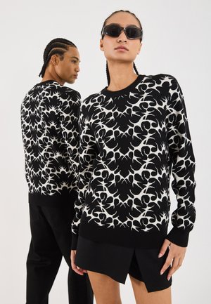 Gebreide trui met een zwart-wit abstract patroon, ronde halslijn en lange mouwen, gecombineerd met zwarte shorts. Glad textuur overal.