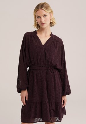 Femme aux cheveux blonds portant une robe longue manches longues violet foncé avec ceinture et boucles d'oreilles créoles dorées sur un fond clair uni.