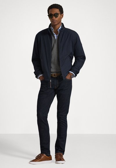 Ναυτική μπomber jacket πάνω από ένα γκρι πουλόβερ, μπλε πουκάμισο, σκούρο τζιν και καφέ αθλητικά παπούτσια με λευκή σόλα. Σκούρα γυαλιά ηλίου ολοκληρώνουν το look.