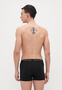 Svarte bomullsbokser med en bred linning som har teksten "COPENHAGEN STUDIOS". Modellen har kort hår og en tatovering på ryggen.