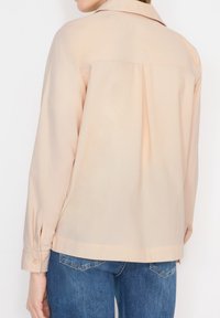 Personne portant une chemise à manches longues beige clair avec un pli au dos, associée à un jean en denim bleu.