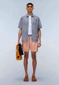 Short-sleeve, navy and white patterned shirt over a white t-shirt, orange patterned shorts, brown slip-on shoes, and yellow sunglasses.