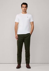 Hvid t-shirt med korte ærmer og sort logo, kombineret med mørkegrønne bukser. Model iført brune støvler, stående foran en neutral baggrund.