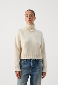 Kremfarget høyhalset genser med ribbestrikket kant og mansjetter, laget av teksturert strikket stoff. Kombinert med lyseblå denimjeans.