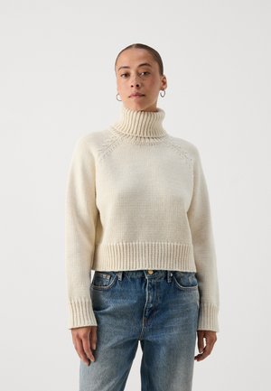 Kreminė turtleneck megztinis su briaunotu apatiniu kraštu ir rankogaliais, pagamintas iš tekstūruoto megztinio audinio. Suderintas su šviesiai mėlynomis džinsinėmis kelnėmis.