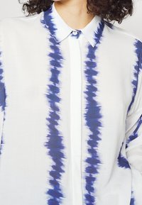 Camicia bianca con strisce tie-dye blu, caratterizzata da un colletto button-down, pattina e un tessuto morbido e leggero.