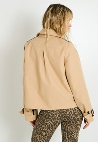 Femme de profil portant une veste cropped beige et un pantalon à imprimé léopard sur fond blanc uni.