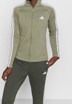 Person iført olivengrøn Adidas lynlåsjakke og matchende bukser med hvide striber ned langs ærmerne og benene, stående mod en ensfarvet baggrund.