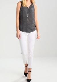 Donna che indossa una blusa senza maniche grigia scura, pantaloni bianchi aderenti e décolleté neri con punta a punta e cinturino alla caviglia, in piedi su uno sfondo neutro.