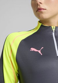 Harmaa urheilupaita, jossa on puoliveto-ominaisuus, fluoresoivan keltaiset yksityiskohdat ja vaaleanpunainen Puma-logo. Teksturoitu materiaali, jossa on erottuvat saumat.