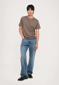 Brown patterned t-shirt, short sleeves, paired with light blue wide-leg jeans. Black shoes complete the outfit against a plain background.