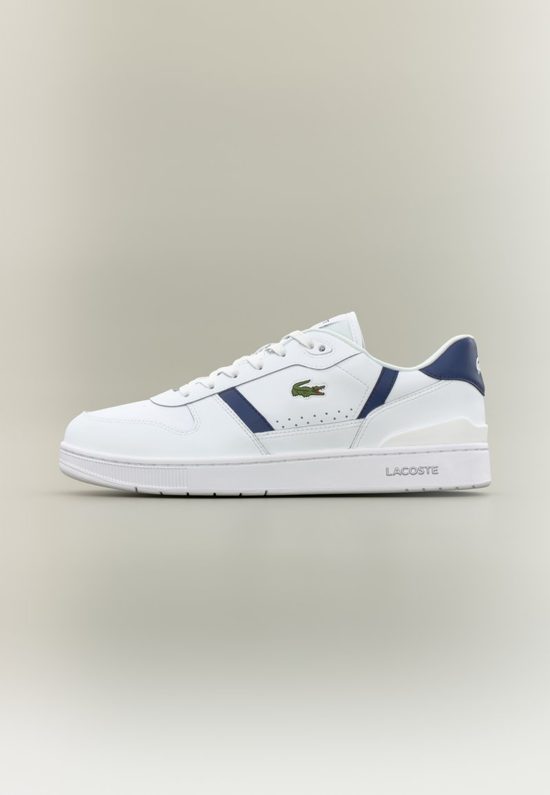 Λευκό sneaker Lacoste με ναυτικό μπλε λεπτομέρειες, πράσινο λογότυπο κροκοδείλου, σχέδιο με κορδόνια και επωνυμία στη σόλα σε ουδέτερο φόντο.