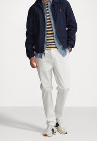 Marineblau leichte Jacke über einem Jeanshemd, gestreiftes gelbes und marineblaues Hemd, weiße Hose und mehrfarbige Sneaker mit strukturiertem Sohlen.