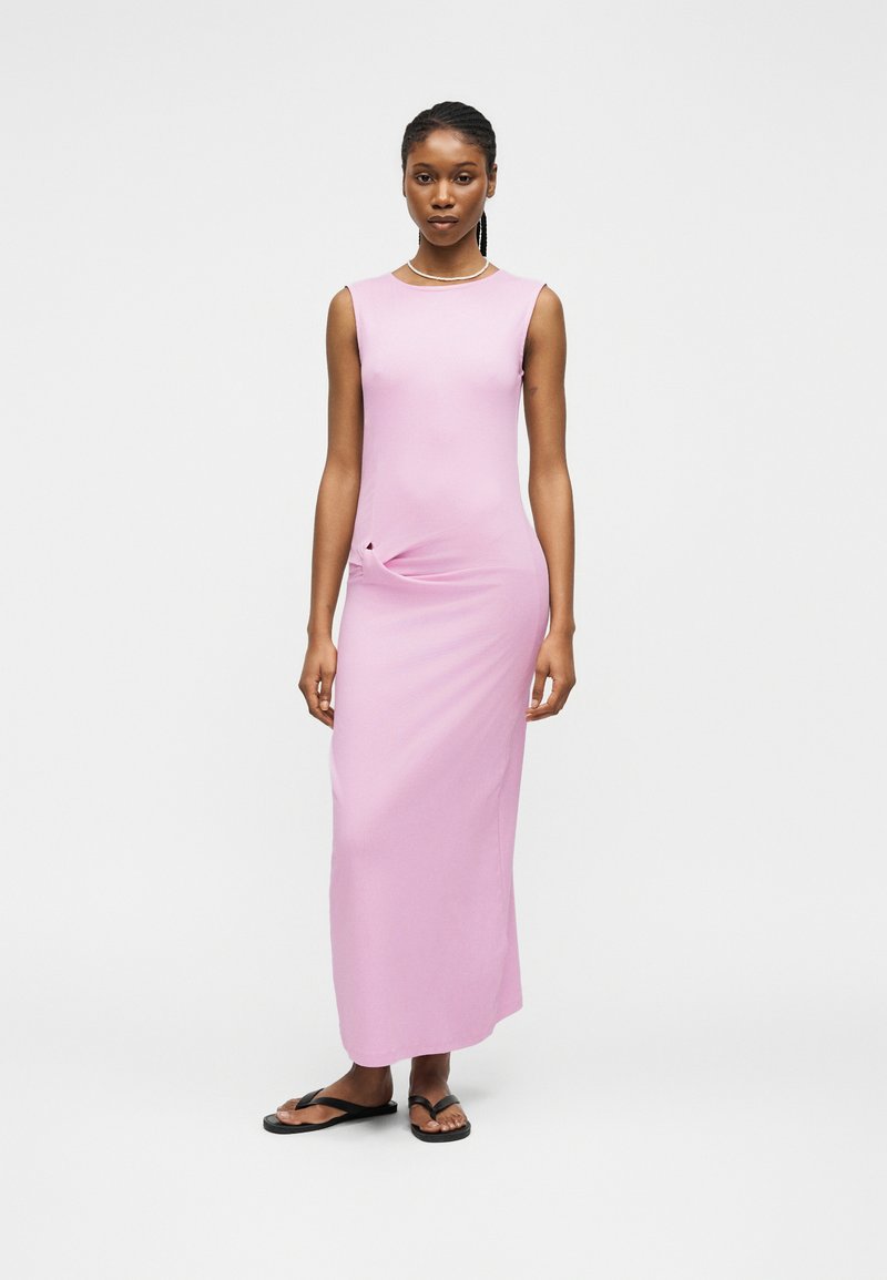 Γυναίκα που στέκεται φορώντας ένα αμάνικο, ανοιχτό μοβ μάξι φόρεμα με λεπτομέρεια σε πλαϊνό γύρισμα και μαύρες σαγιονάρες μπροστά σε απλό φόντο.