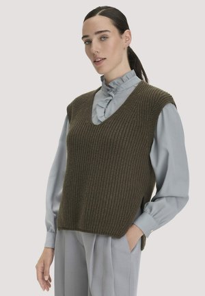 Frau mit dunkel gebundenen Haaren, die eine graue Bluse mit Rüschen und eine olivgrüne gestrickte ärmellose Weste trägt, steht mit einer Hand in der Tasche.