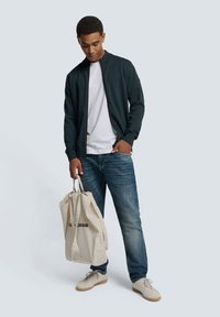 Donkergroene bomberjack, witte t-shirt, blauwe jeans en beige canvas draagtas. Casual schoenen met een lichte kleurenschema.