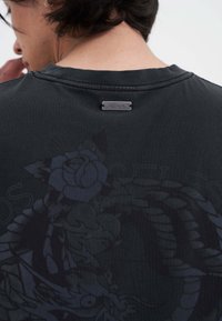 Czarny bawełniany t-shirt z subtelnym motywem kwiatowym i grafiką smoka, z metaliczną metką marki przy dekolcie oraz klasycznym okrągłym dekoltem.