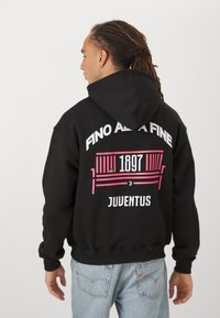 Čierna mikina s kapucňou vyrobená z mäkkej tkaniny. Zadná strana obsahuje ružovú a bielu grafiku: "FINO AL FINE," "1897," a "JUVENTUS," s dizajnom v tvare pruhu.