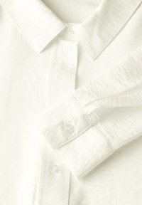 Camicia bianca con colletto classico, chiusura a bottoni e maniche lunghe. Il tessuto è liscio, con una leggera lucentezza e una consistenza morbida.