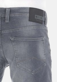 Tył szarych dżinsów pokazujący prawą tylną kieszeń z haftowanym wzorem oraz czarną etykietą "Mustang True Denim" na pasku.