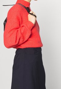 Persoon in een felrode gebreide trui en een donkere high-waisted rok, met een zwarte band over de schouder, tegen een eenvoudige achtergrond.